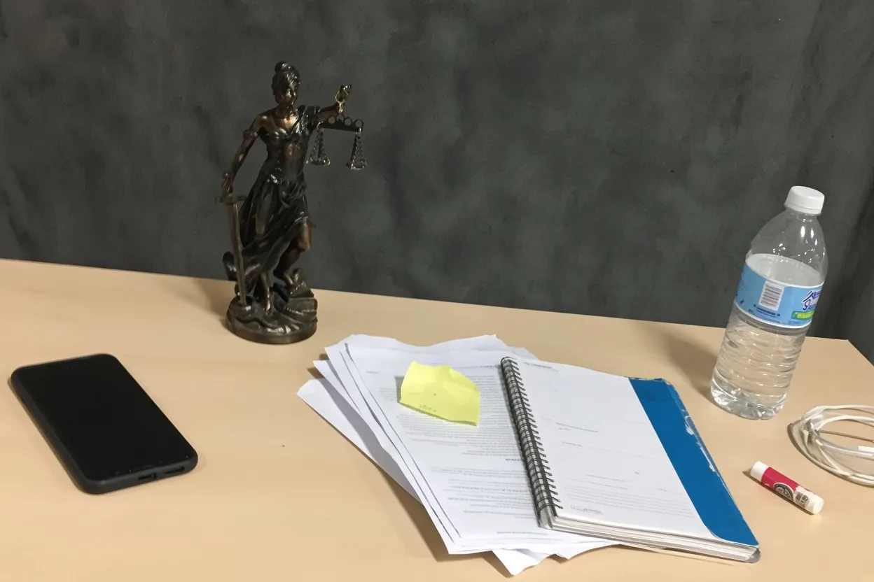A bronze Lady Justice statue on a wooden desk beside a notebook, magnifying glass, and clipboard