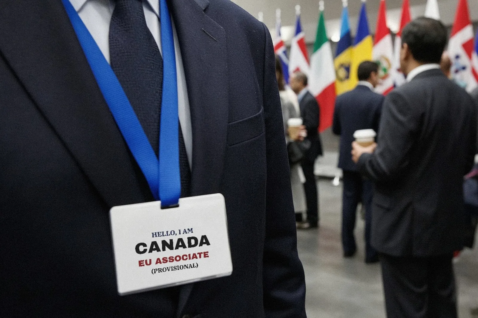 Close view of a man’s navy suit jacket with a blue lanyard and a white name badge reading “Hello, I am Canada. EU Associate (Provisional),” with people in suits and a row of national flags softly behi