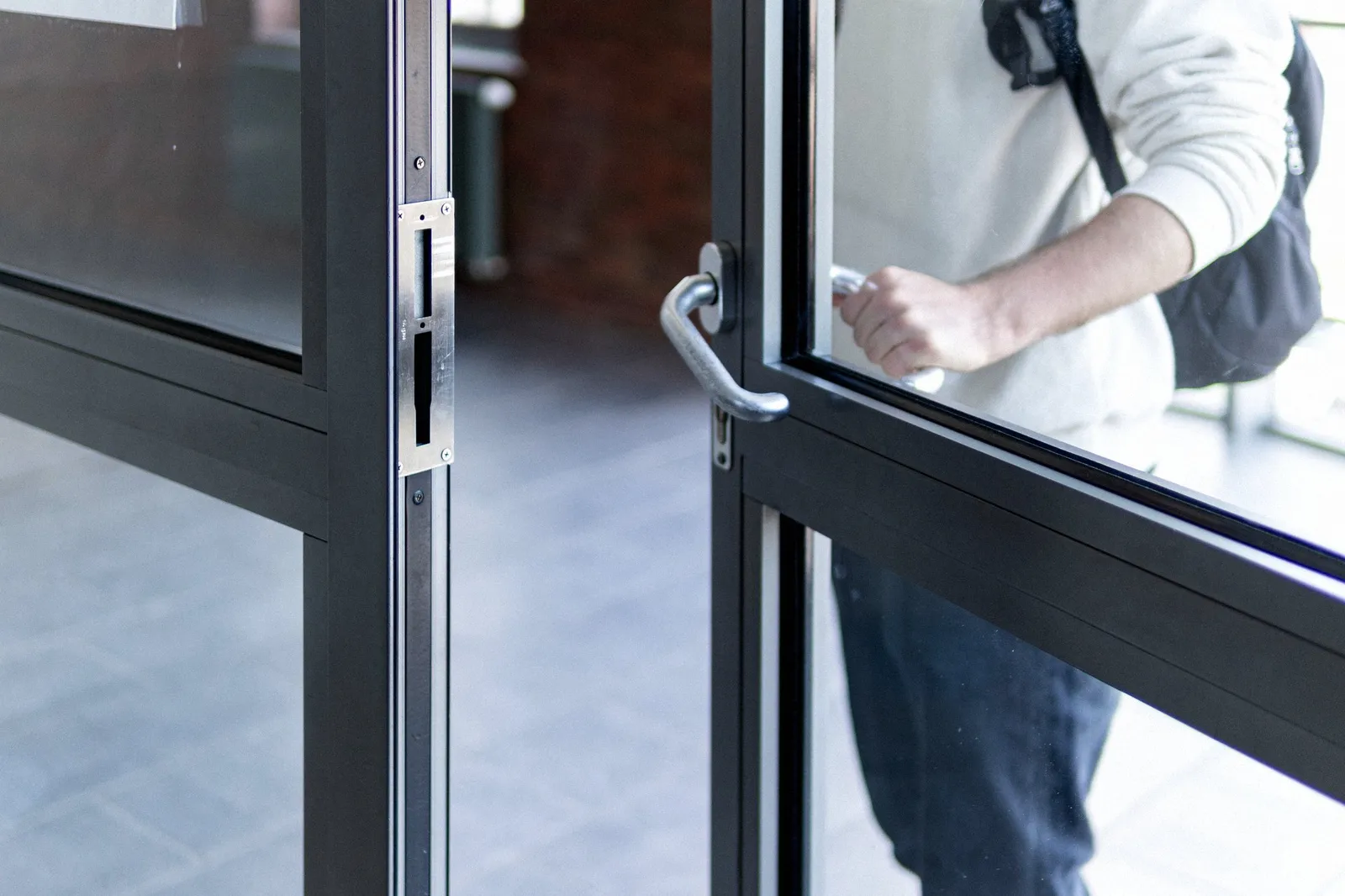 A man in a light sweater with a backpack holds open a glass door by its metal handle, seen from the chest down.