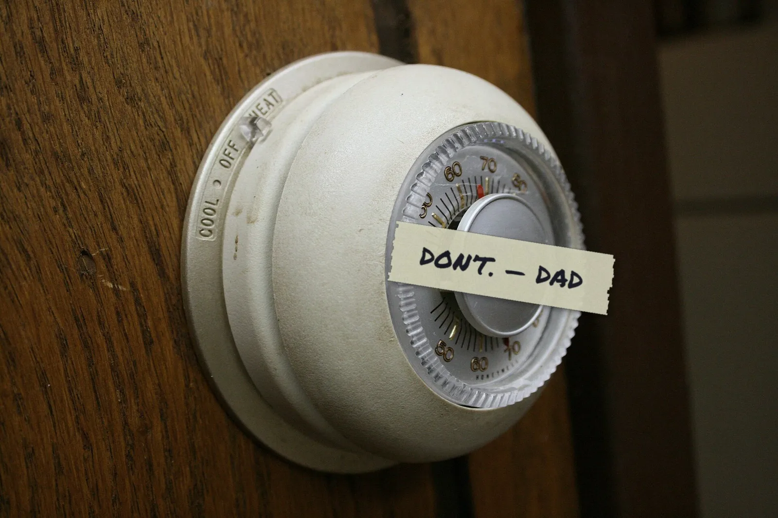 An old round dial thermostat on a wood-paneled wall with a strip of masking tape across it reading Don't, Dad