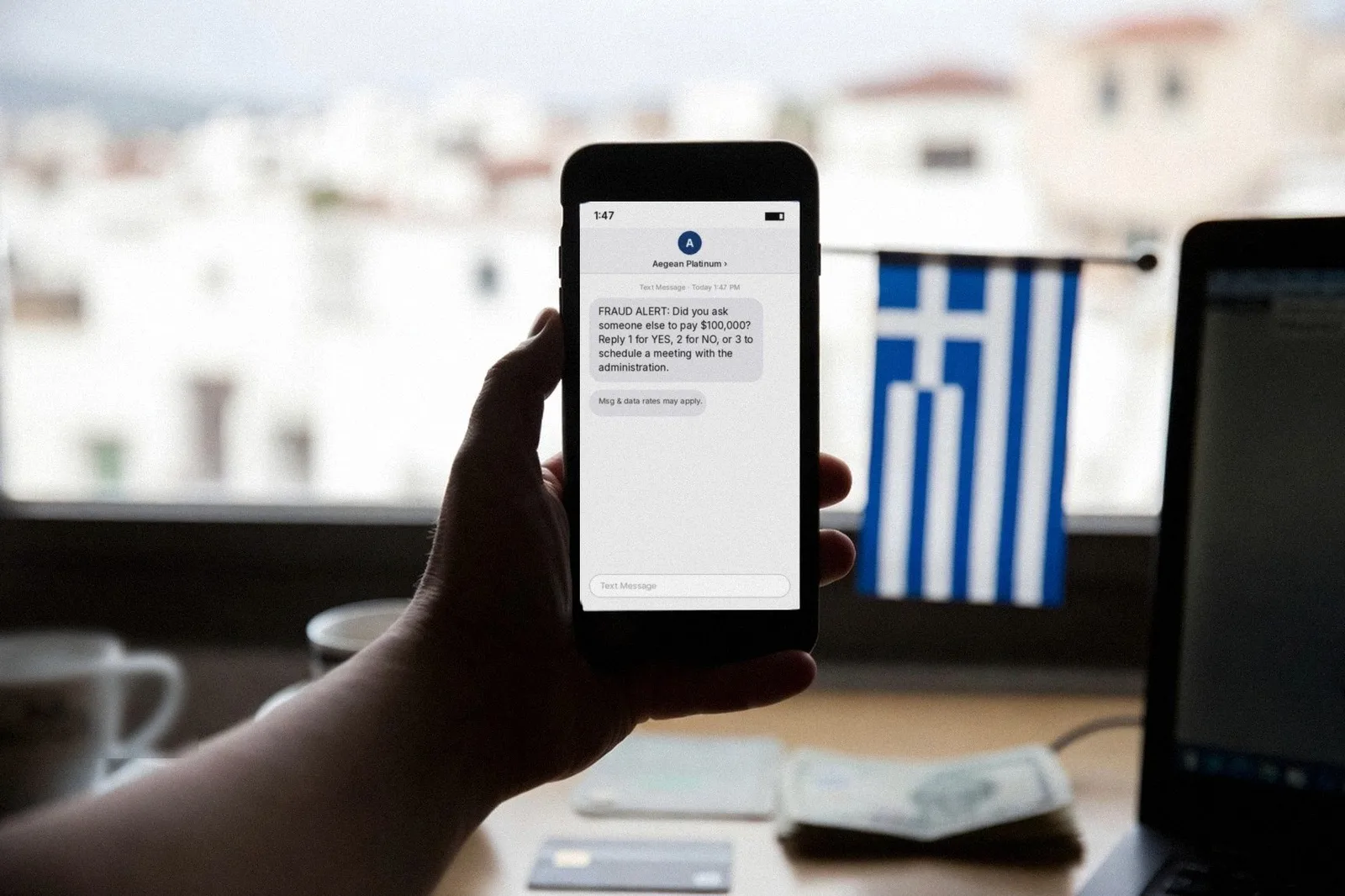 A hand holds up a smartphone showing a bank fraud alert text, above a desk with a credit card, a paper bill and a small Greek flag