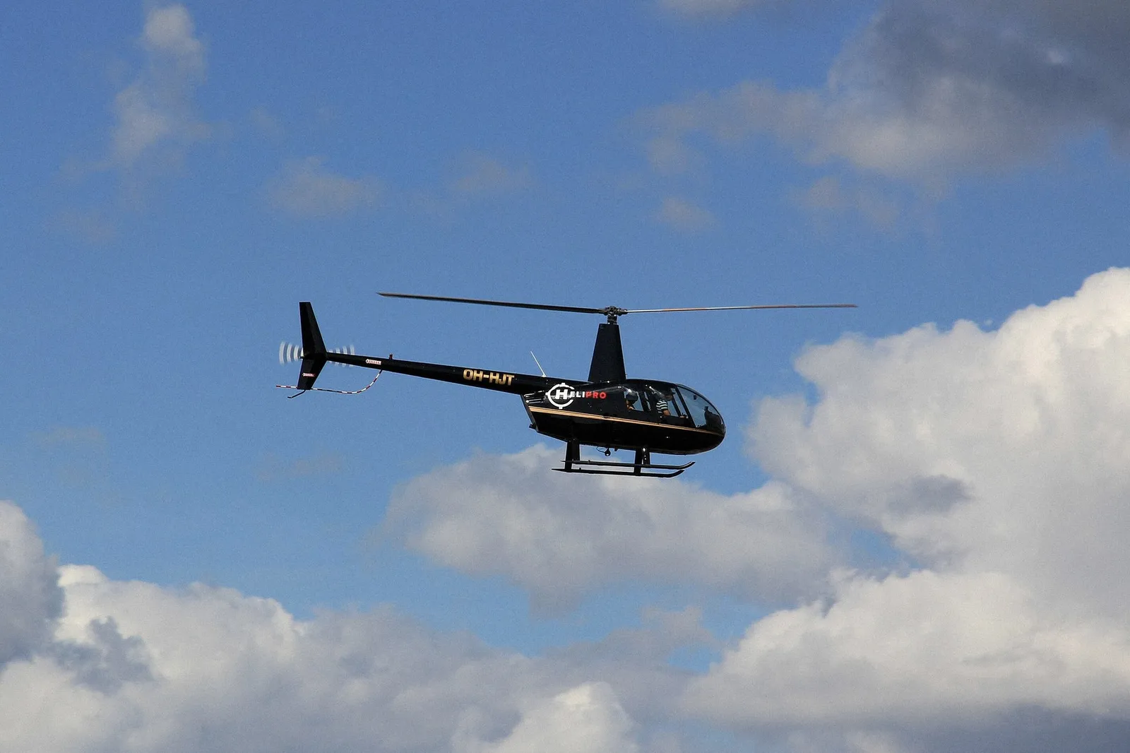 A dark Robinson R44 helicopter flying against a cloudy sky