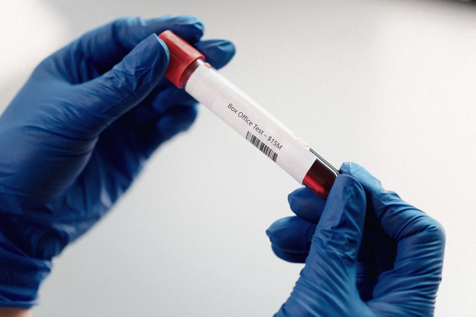 Two hands in blue gloves hold a red-capped sample tube labeled “Box Office Test – $15M” with a barcode
