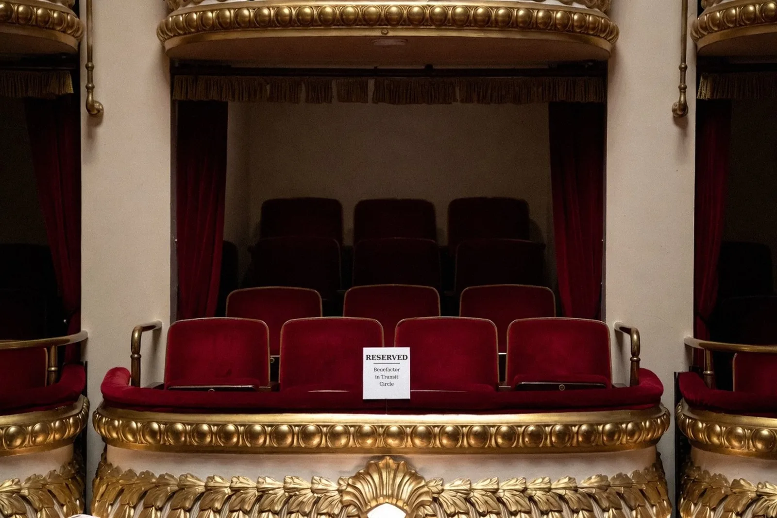 An empty private theater box with red velvet seats and a gold railing, with a small white card on the ledge reading Reserved, Benefactor in Transit Circle.