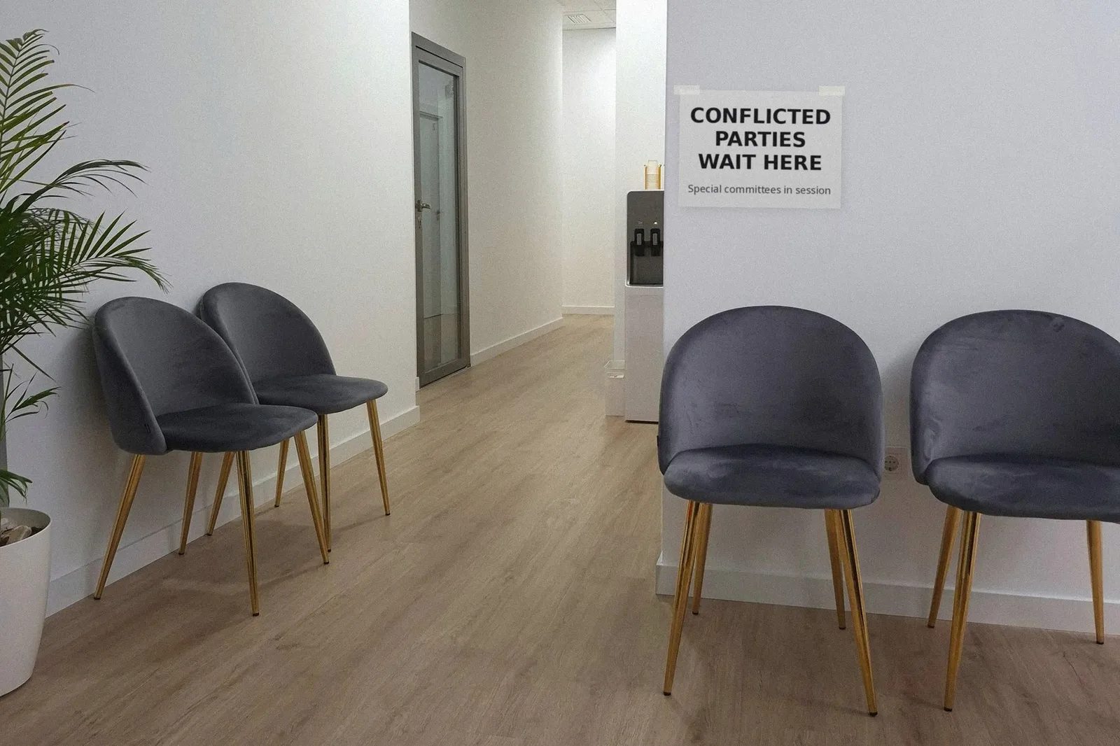 An office hallway with grey velvet chairs, a water cooler and a paper sign taped to the wall reading Conflicted Parties Wait Here, Special committees in session
