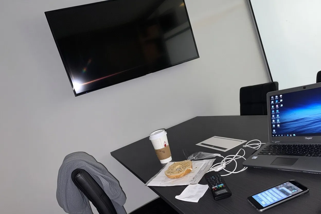 An empty meeting room with a wall-mounted television above a dark conference table and black office chairs