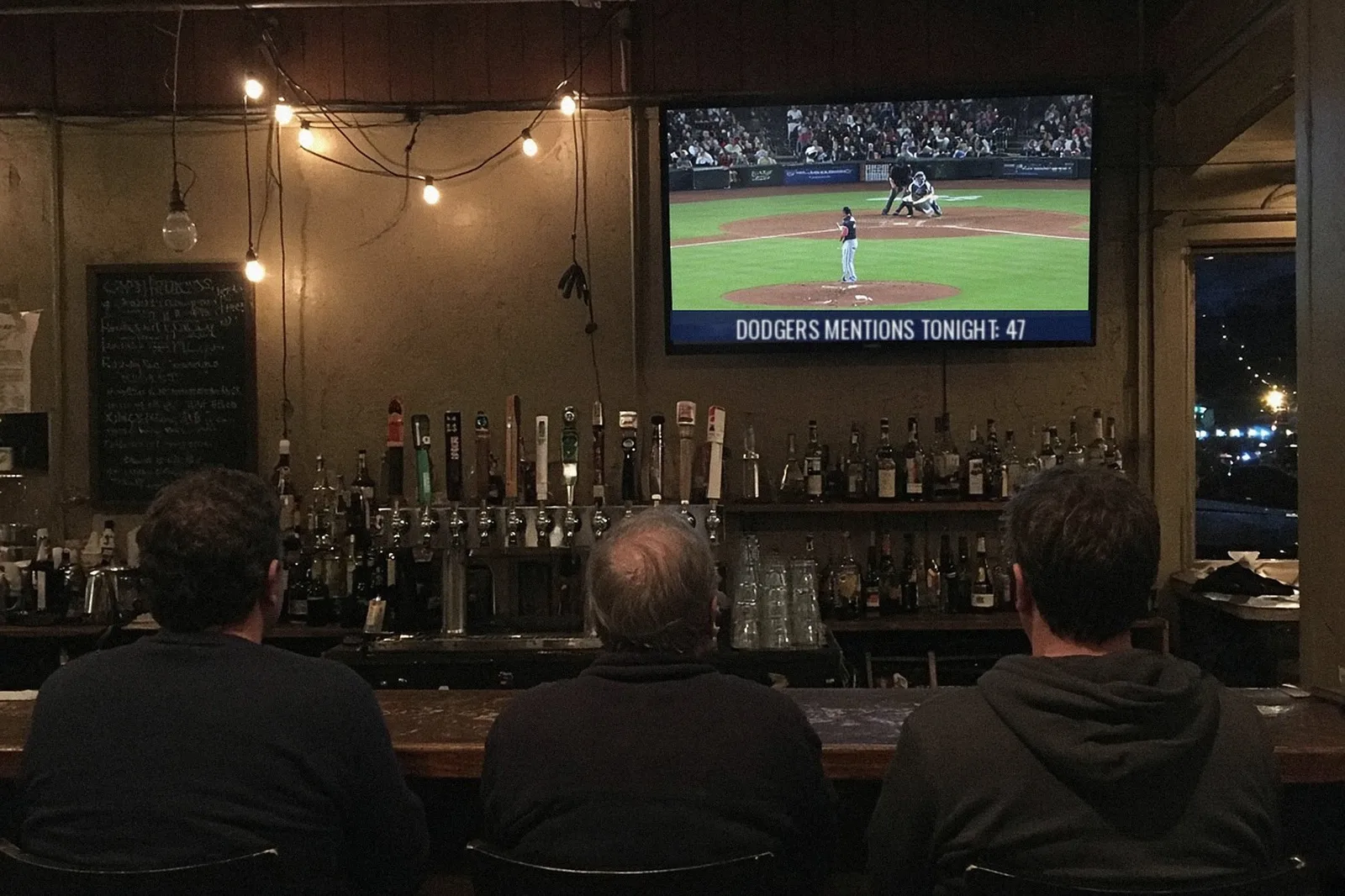 Bar patrons watch a baseball game on a TV whose on-screen banner reads “Dodgers mentions tonight: 47.”