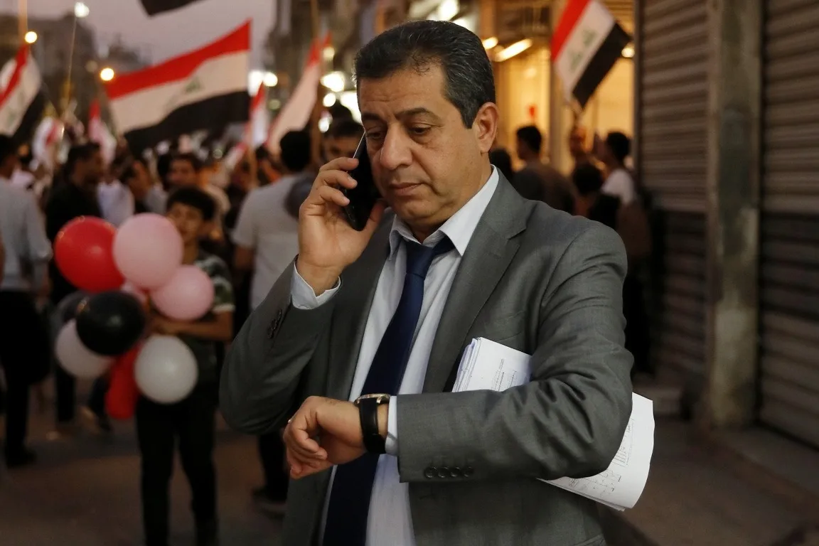 Iraqi official in a gray suit talks on his phone and checks his watch as families celebrate with flags and balloons on a Baghdad street at dusk