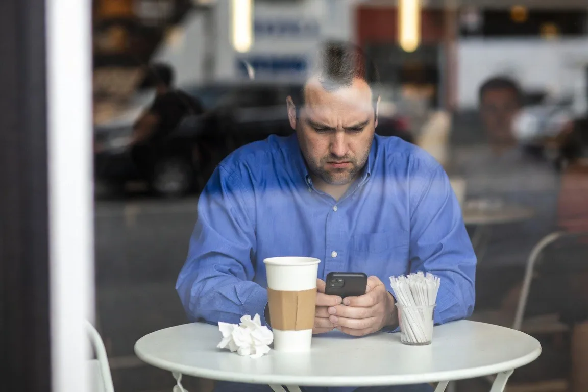 Frustrated man in a blue shirt reads his phone at a coffee shop table.