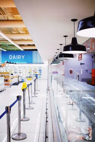An empty, brightly lit supermarket deli counter and dairy aisle with stanchions and a yellow wet-floor sign