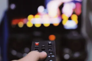 A hand holding a TV remote in front of a blurred television