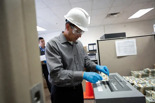 An office worker in a white hard hat, safety goggles and blue gloves carefully feeds a hundred-dollar bill into a paper shredder beside stacks of banded cash and an orange traffic cone.