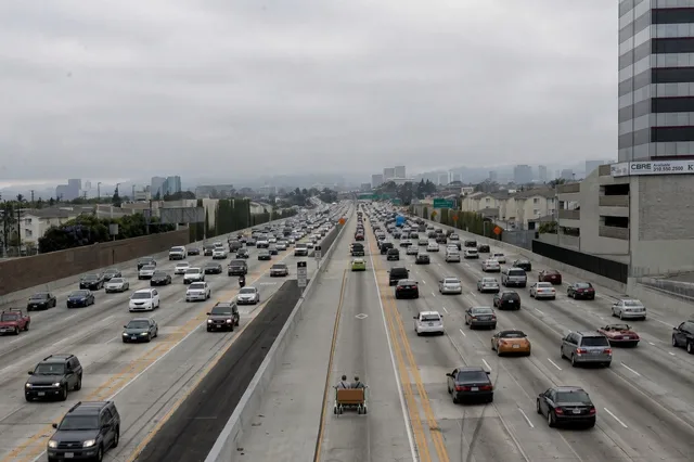 Photo illustration of two men seen from behind riding a tan love seat mounted on two electric rental scooters down a freeway carpool lane beside heavy traffic.