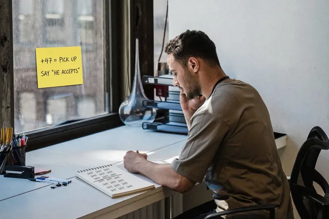 A young man on the phone beside a window sticky note reading +47 = pick up, say he accepts