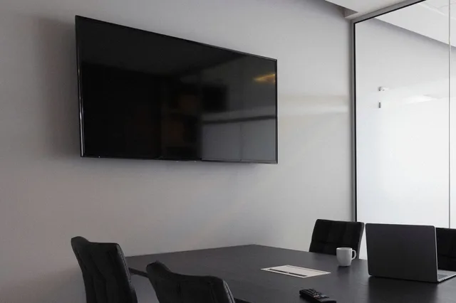 An empty meeting room with a wall-mounted television above a dark conference table and black office chairs
