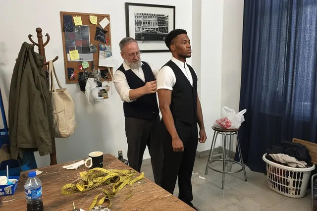 A tailor in a suit shop measures a man in a black vest and white shirt, surrounded by suits on mannequins