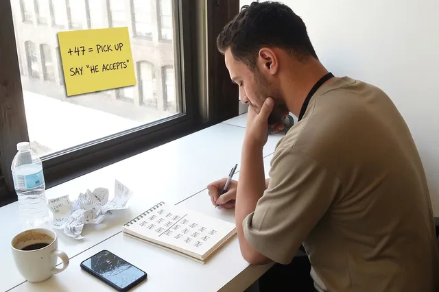 A young man on the phone beside a window sticky note reading +47 = pick up, say he accepts