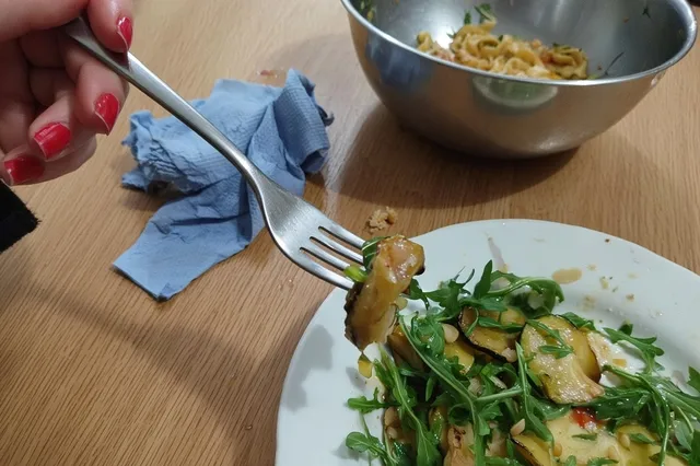 A woman’s hand reaches across a wooden restaurant table with a fork toward a plate of pasta in front of another diner.