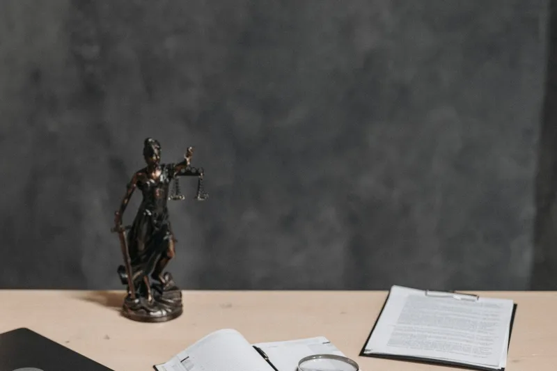 A bronze Lady Justice statue on a wooden desk beside a notebook, magnifying glass, and clipboard