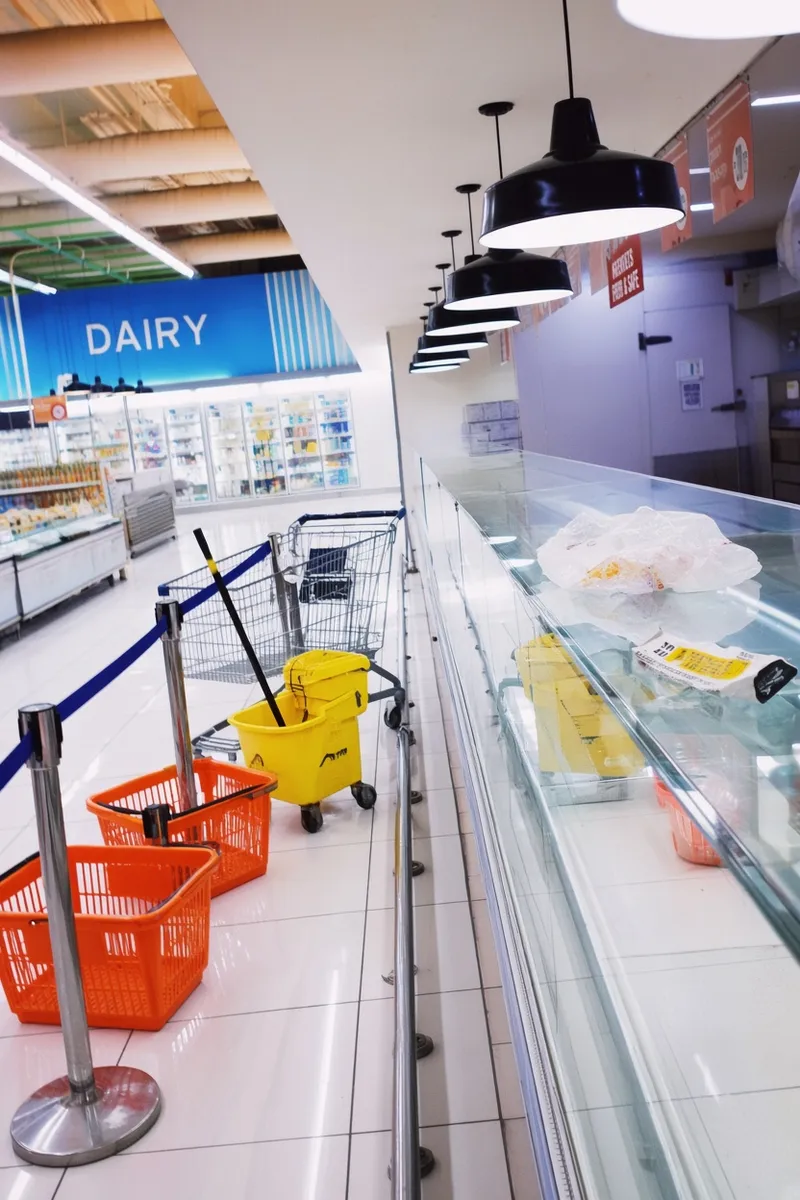 An empty, brightly lit supermarket deli counter and dairy aisle with stanchions and a yellow wet-floor sign