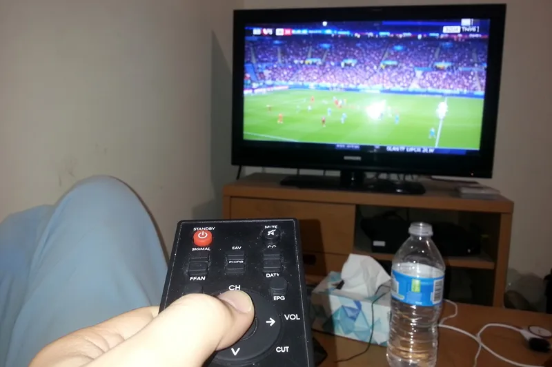 A hand holding a TV remote in front of a blurred television