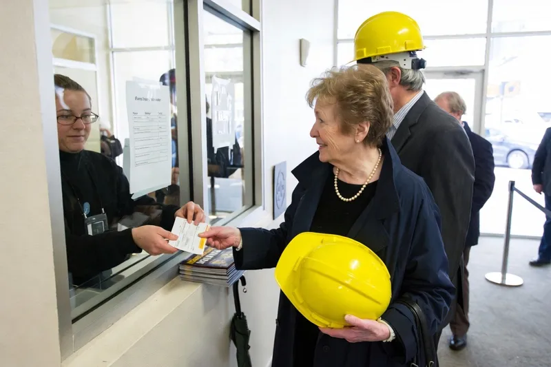 A woman in pearls holds a yellow hard hat while buying tickets at a box office window