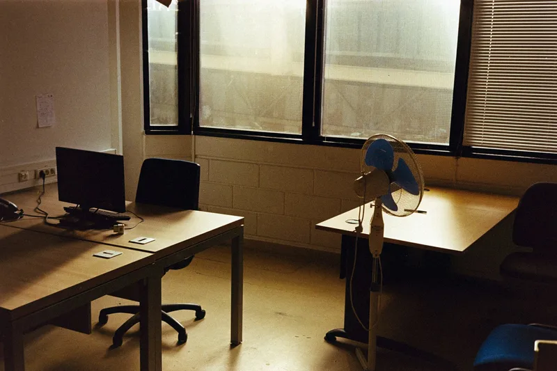 An empty office with wooden desks, a computer workstation, and a blue standing fan by the windows
