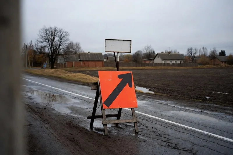 An orange detour sign with a black arrow on a wooden sawhorse points off a wet rural road toward a muddy field, with a blank sign and low brick houses behind it.