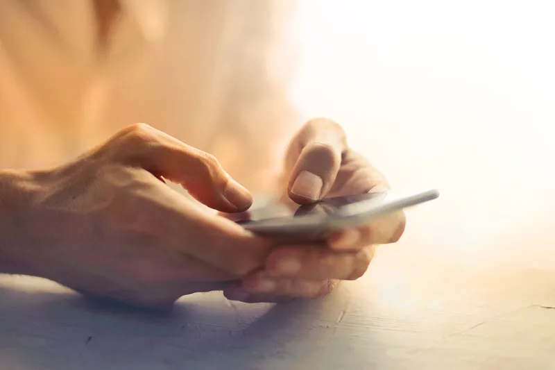Close-up of two hands holding and tapping a smartphone on a wooden table in warm light