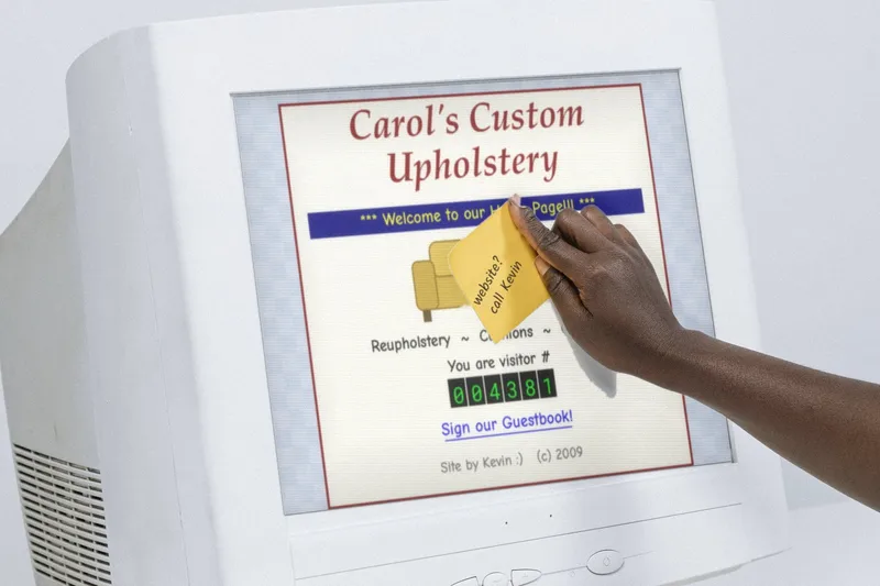 A hand pressing a yellow sticky note that reads “website? call Kevin” onto an old beige CRT monitor showing a homemade upholstery shop homepage with a visitor counter