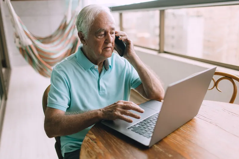 Older man at a wooden table with a laptop, holding a phone to his ear and waiting