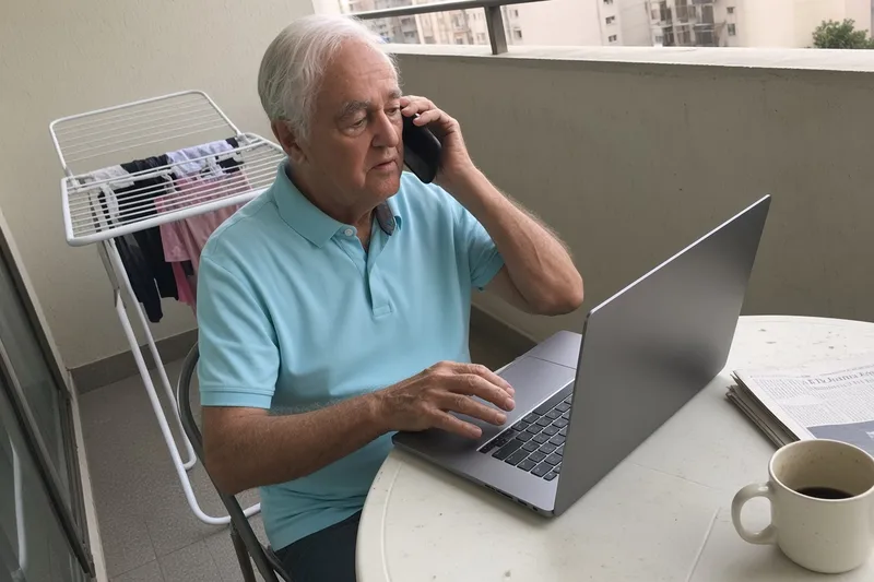 Older man at a wooden table with a laptop, holding a phone to his ear and waiting