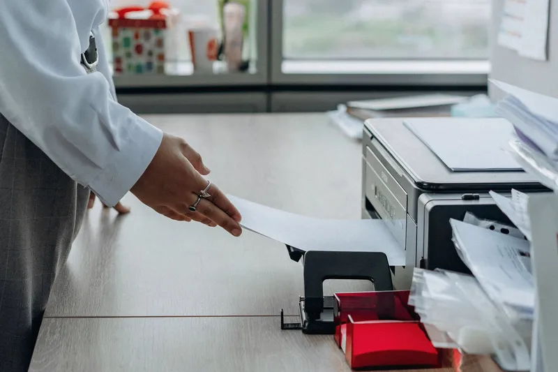 A woman in a white blouse reaches for a sheet of paper coming out of an office printer on a wooden desk by a window