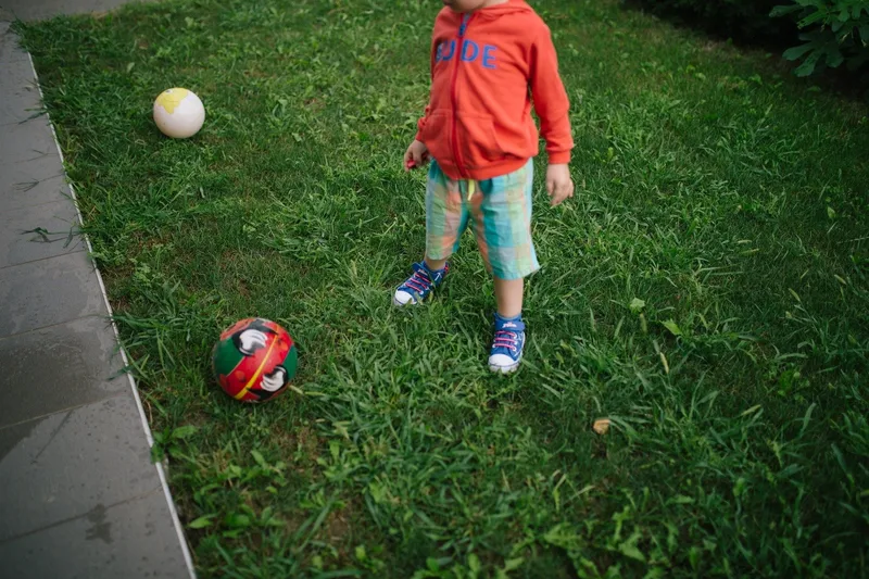 A small child in a red hoodie, plaid shorts and sneakers stands on a backyard lawn beside two balls, face out of frame.